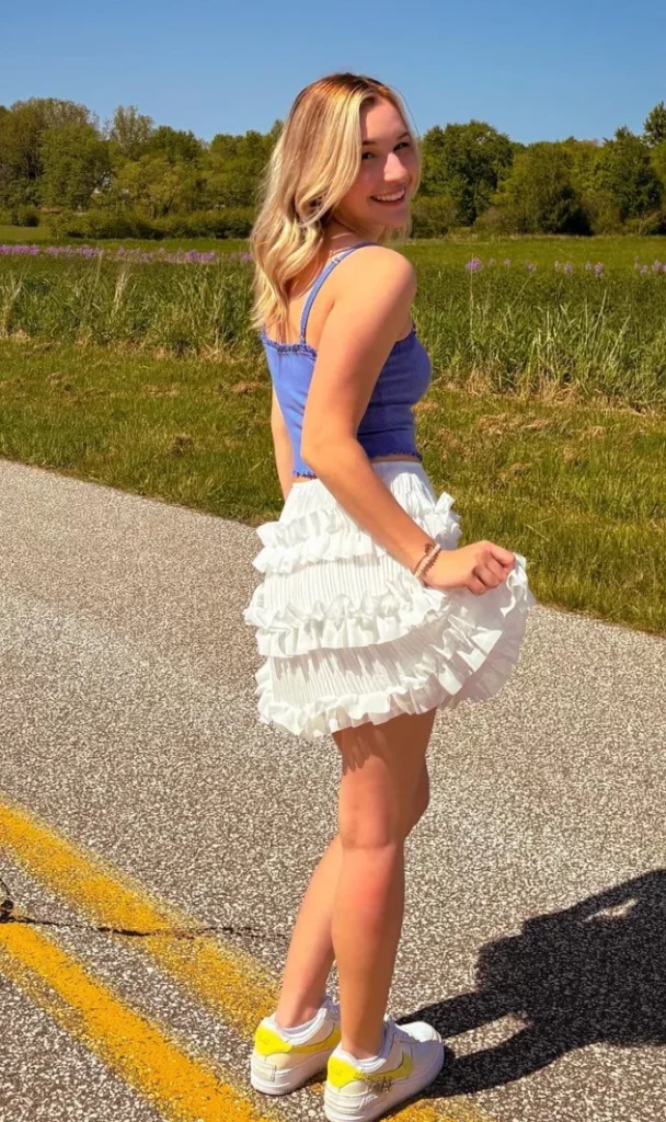 Skirt Outfits - Blonde woman outdoors wearing a white ruffled mini skirt paired with a blue tank top and white sneakers, smiling and looking back over her shoulder on a sunny day.