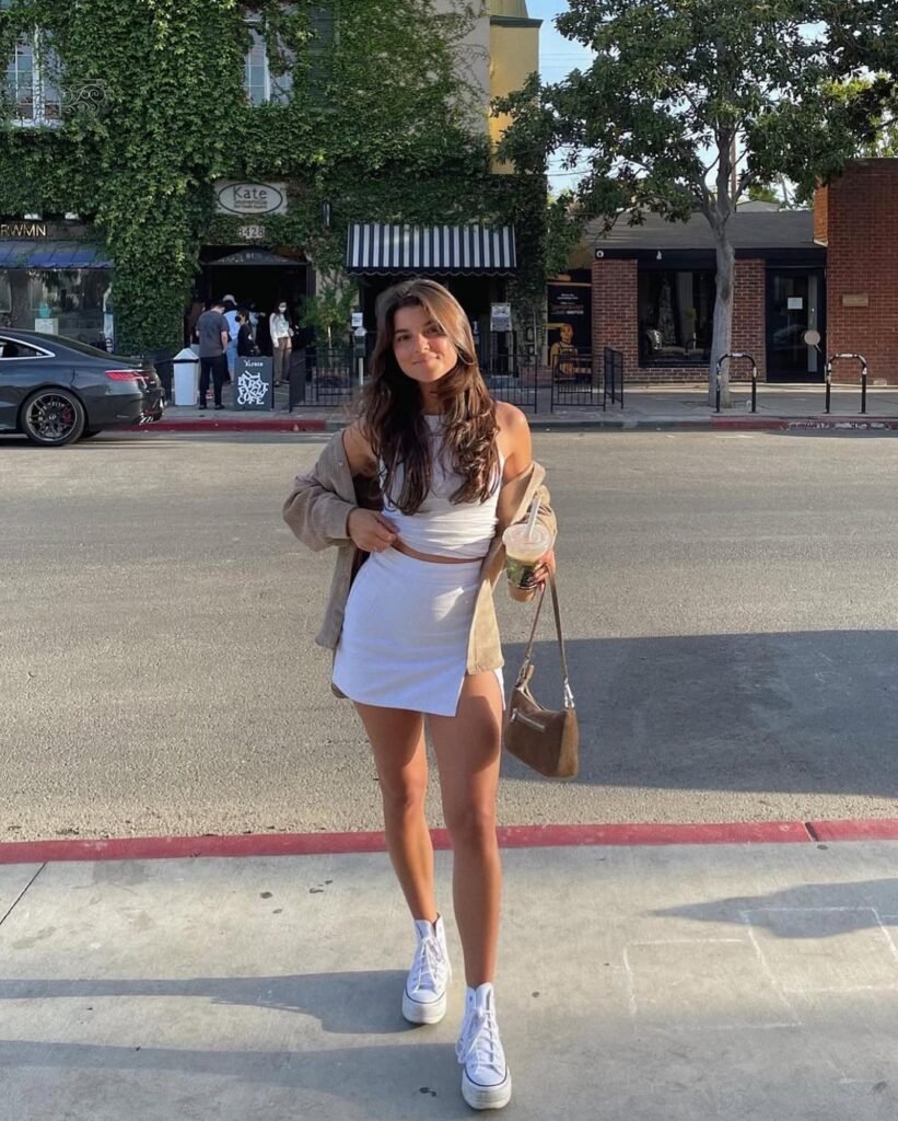 A young woman wearing a white crop top and white asymmetrical mini skirt with a beige overshirt and platform white sneakers, standing on a city street — Skirt Outfits for casual summer city walks.