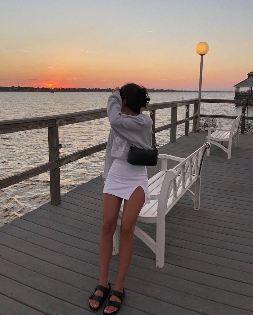  A woman standing on a lakeside boardwalk at sunset, wearing a white mini skirt with a thigh slit and an oversized grey sweatshirt, accessorized with a black shoulder bag and sandals — Cozy Skirt Outfits for sunset by the water.

