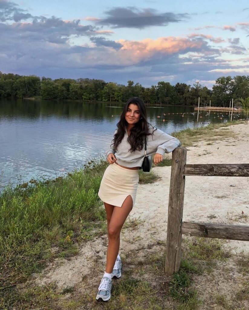 A woman in a beige mini skirt with a front slit, paired with a grey sweatshirt and running shoes, posing by a lakeside fence — Sporty and cute Skirt Outfits for outdoor adventures.
