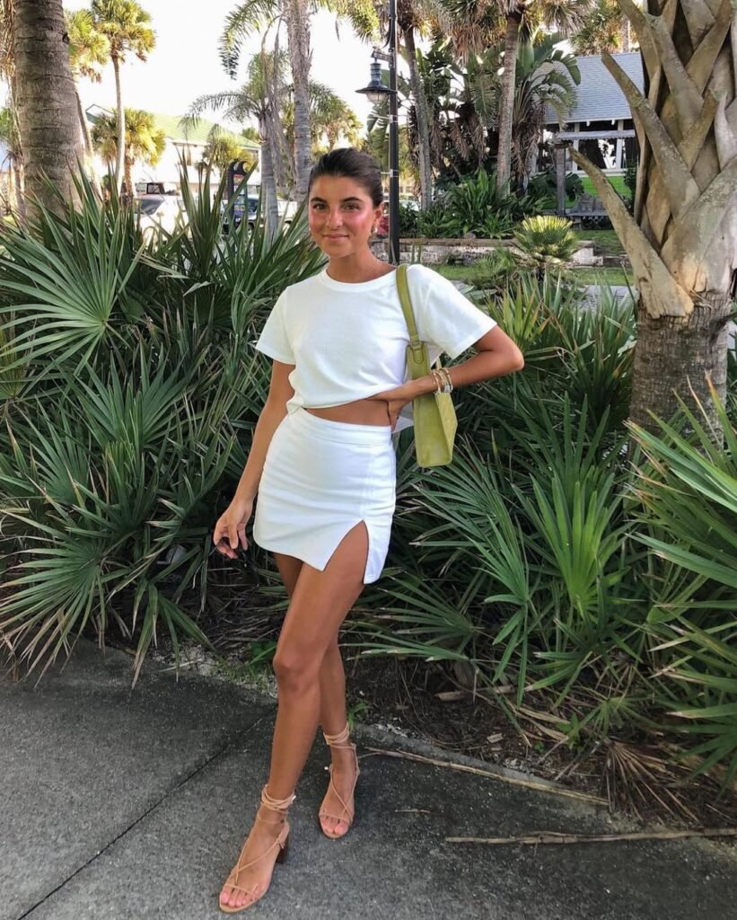 A woman wearing a coordinated all-white outfit with a crop top and high-slit mini skirt, accessorized with nude block heels and a green shoulder bag, standing among palm trees — Elegant Skirt Outfits for tropical getaways.