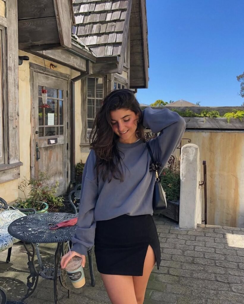 A woman in a black mini skirt and oversized grey sweatshirt, holding a coffee while smiling outdoors in a rustic setting — Cozy and cute Skirt Outfits for cooler mornings.