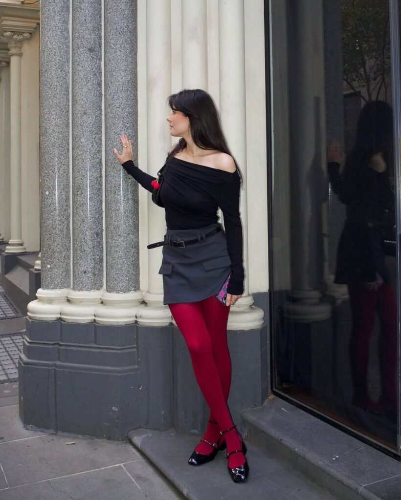 Woman dressed in an off-shoulder black top and a grey skirt outfit with red tights outside.