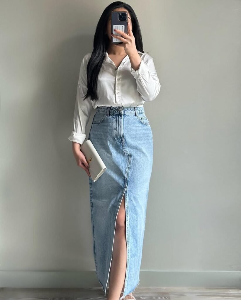 A woman posing indoors in a classic white button-up shirt tucked into a high-slit denim skirt, holding a white clutch — Polished Skirt Outfits perfect for smart-casual looks.