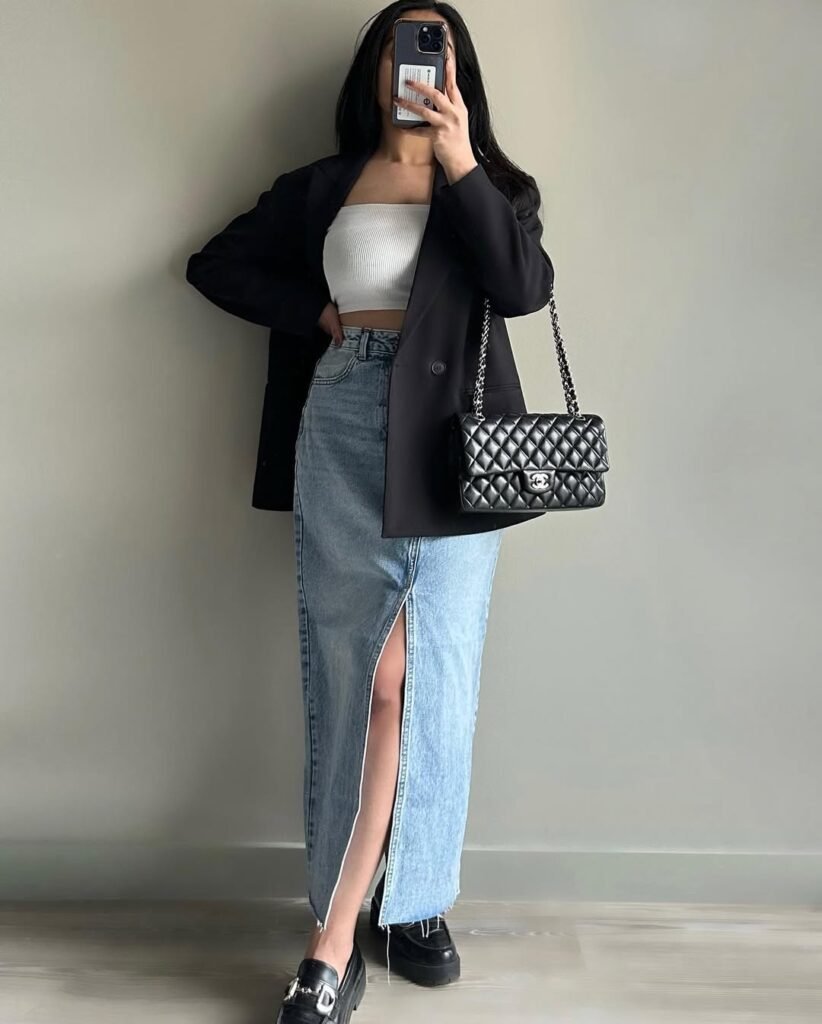 A fashionable woman wearing a white bandeau top, black blazer, and high-slit denim skirt, completed with loafers and a black quilted chain bag — Elevated Skirt Outfits for chic city fashion.