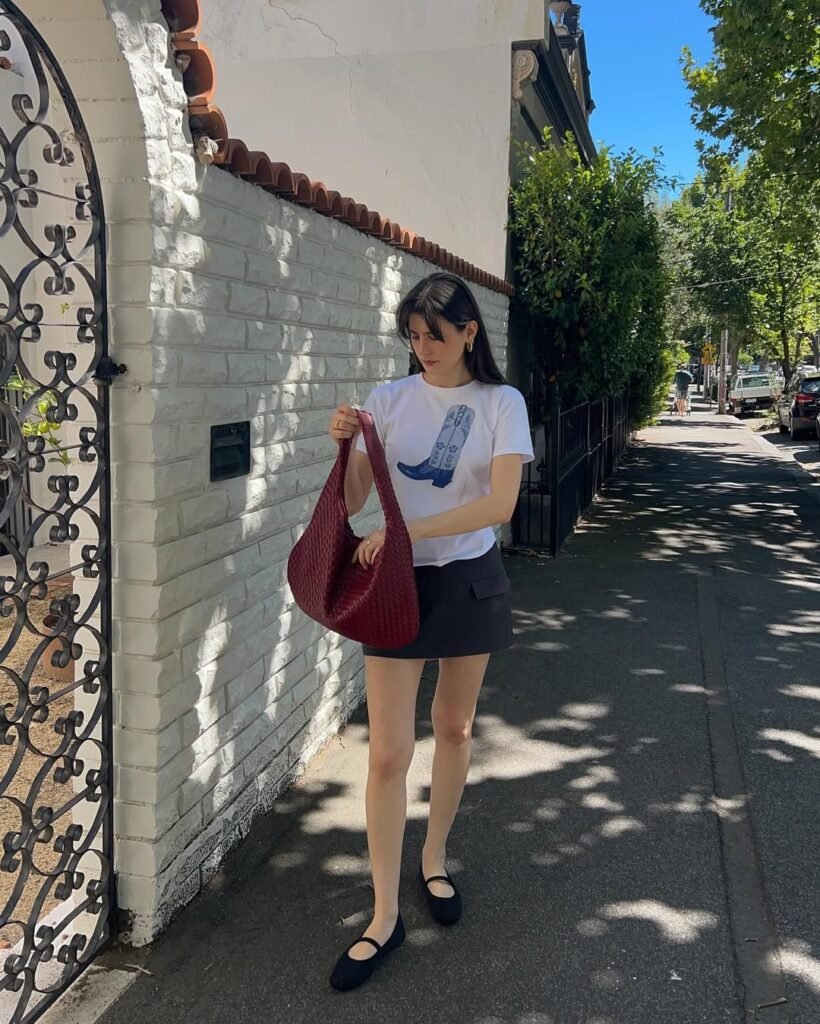 Woman wearing a red crop top and white skirt outfit posing by a wooden fence.