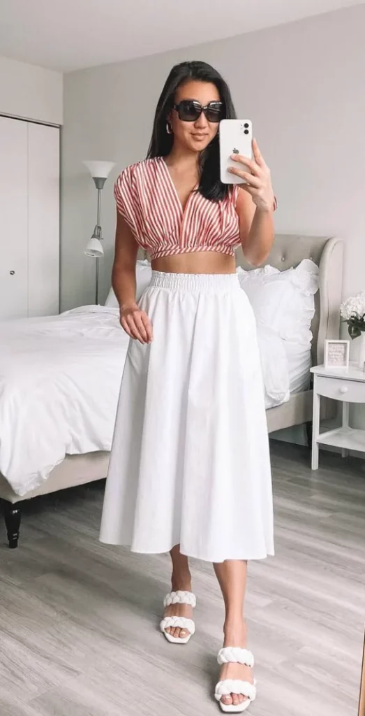 A woman taking a mirror selfie wearing a striped crop top paired with a white midi skirt and white braided sandals, showcasing a stylish and polished skirt outfit perfect for casual and semi-formal occasions.