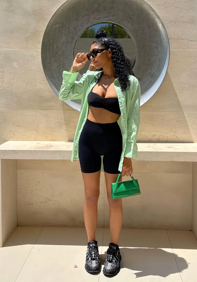 Trendy woman in a black bandeau top and black biker shorts outfit, styled with a light green shirt and mini green handbag.
