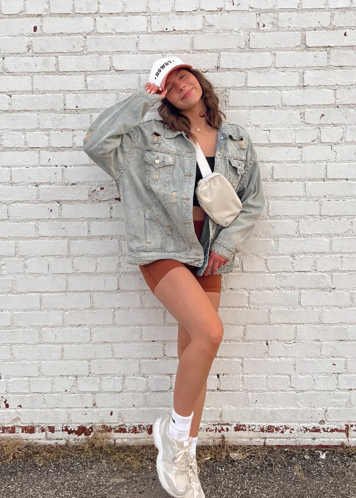 Casual outfit with a gray oversized denim jacket, rust-colored biker shorts, white sneakers, and a crossbody bag, paired with a white cap.