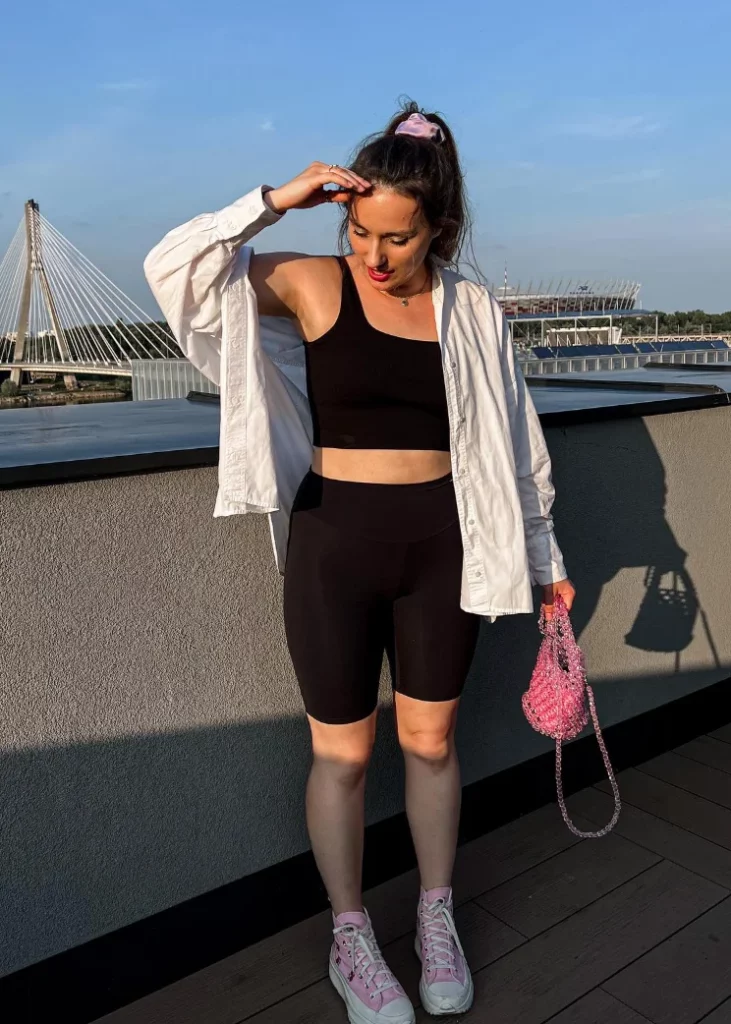 Woman wearing a black crop top and black biker shorts outfit, layered with a white shirt, on a rooftop with a bridge in the background.
