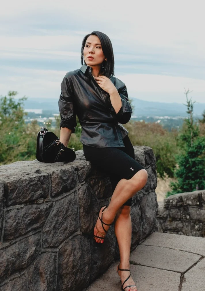 Stylish woman posing in a sleek black leather top and black biker shorts outfit, seated on a stone ledge with scenic nature in the background.