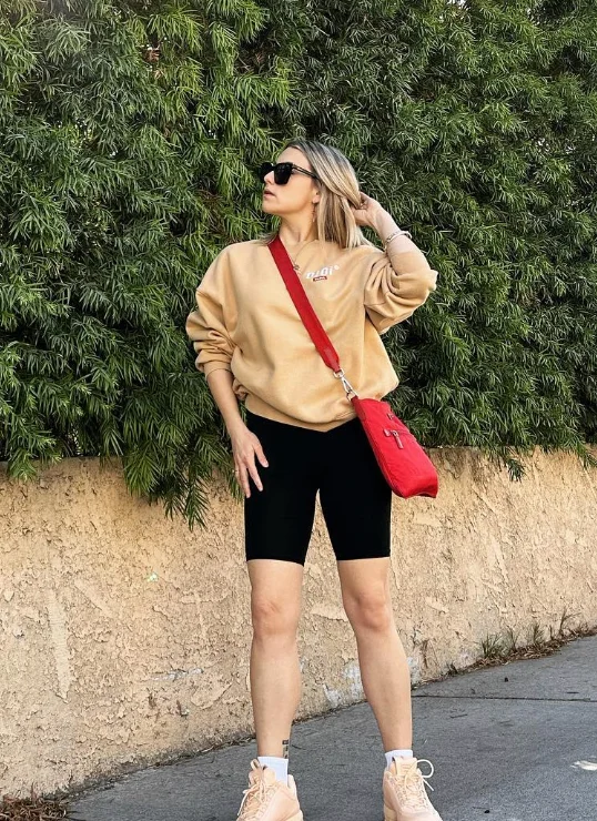 Woman in a beige sweatshirt and black biker shorts outfit accessorized with a red crossbody bag, posing by a leafy green wall.