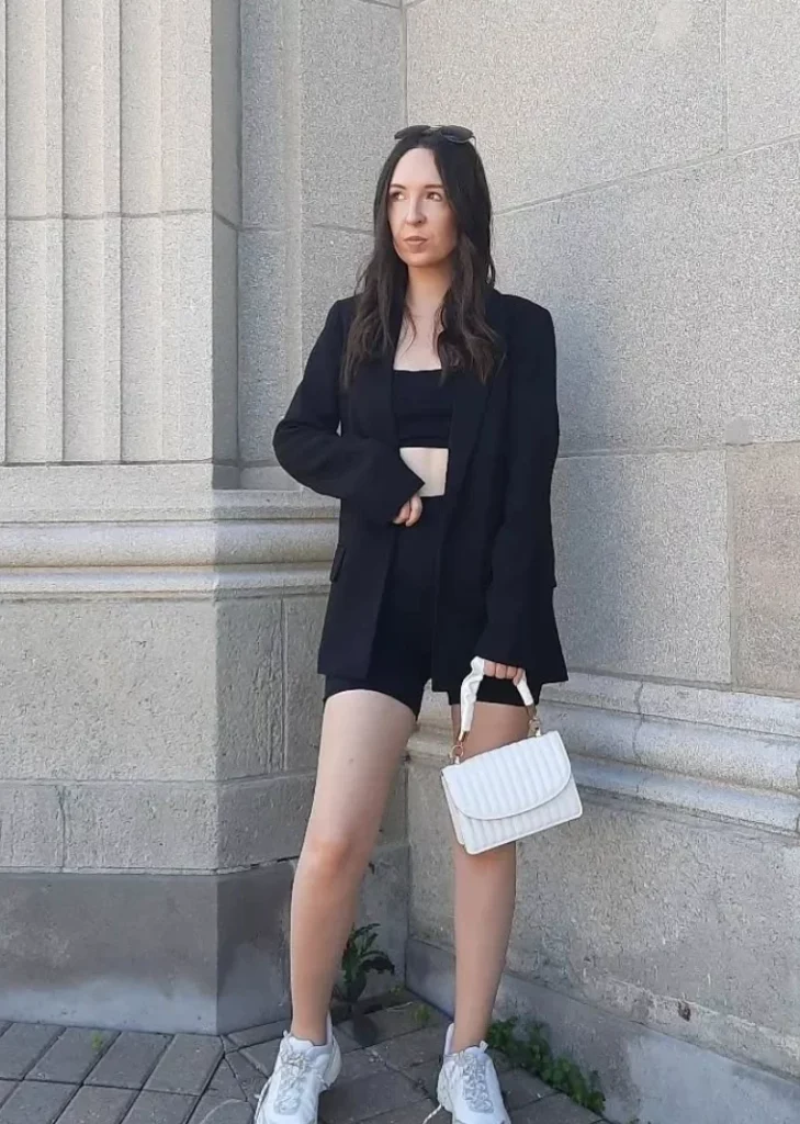 Monochrome look with a black oversized blazer, black crop top, and black biker shorts outfit, finished with white sneakers and a white handbag.