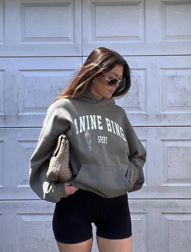 Laid-back woman in a gray “ANINE BING” hoodie and black biker shorts outfit, holding a textured clutch in front of a garage door.