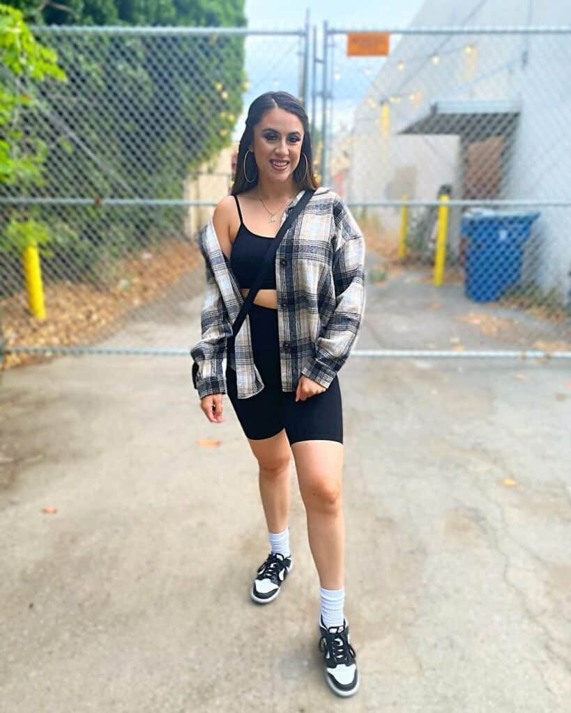 A woman smiling and standing near a fence, wearing black biker shorts, a black tank top, a plaid shirt, white socks, and black and white sneakers – Biker Shorts outfits.