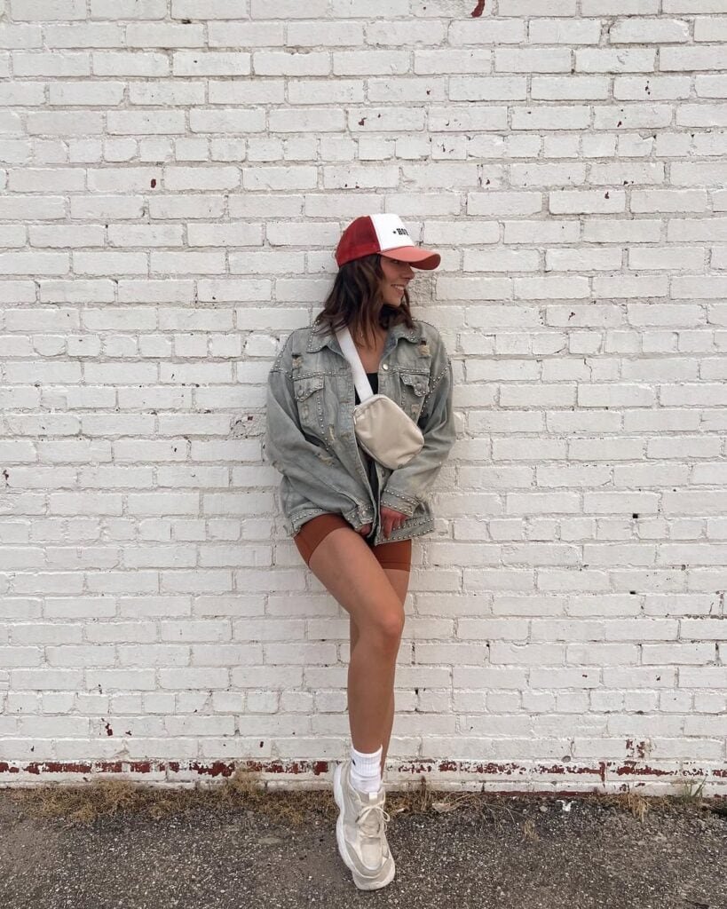 Woman wearing rust-colored biker shorts paired with oversized gray denim jacket, white socks, white chunky sneakers, and a white crossbody bag, posing casually against a white brick wall, showcasing a trendy biker shorts outfit.