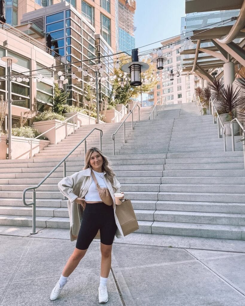 Woman in black biker shorts wearing a white crop top and beige jacket, accessorized with a tan crossbody bag, standing near outdoor stairs with a shopping bag, illustrating a casual and trendy biker shorts outfit.