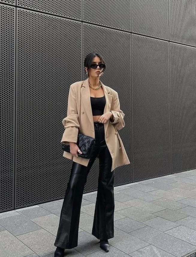 Woman in a camel blazer over a black crop top and black flared leather pants, holding a black clutch, standing in front of a modern building. Blazer Outfits.