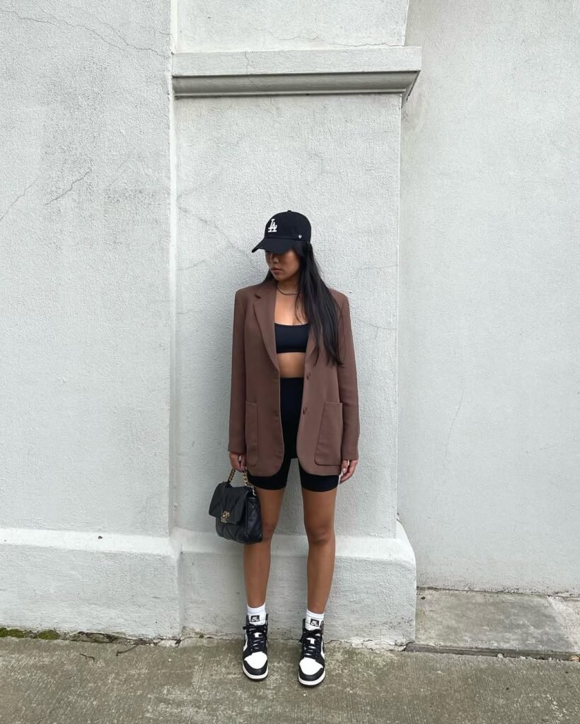 oung woman wearing black biker shorts paired with a brown blazer, black crop top, black and white sneakers, and a black baseball cap, standing against a white textured wall, Biker Shorts outfits.