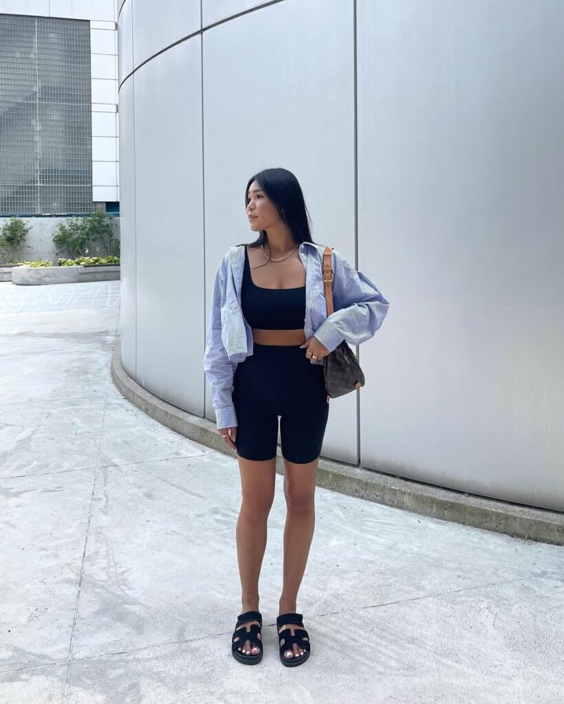 A woman wearing black biker shorts and a black crop top, paired with a light blue oversized striped shirt. She accessorizes with a brown leather handbag and gold jewelry, standing against a modern urban background. This stylish Biker Shorts outfit balances casual and chic vibes.