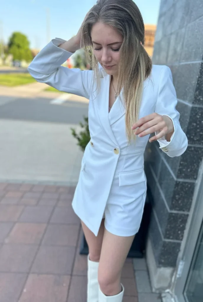 Woman dressed in a white blazer dress with matching boots, outdoors near a sidewalk, looking down with one hand in her hair. Blazer Outfits.