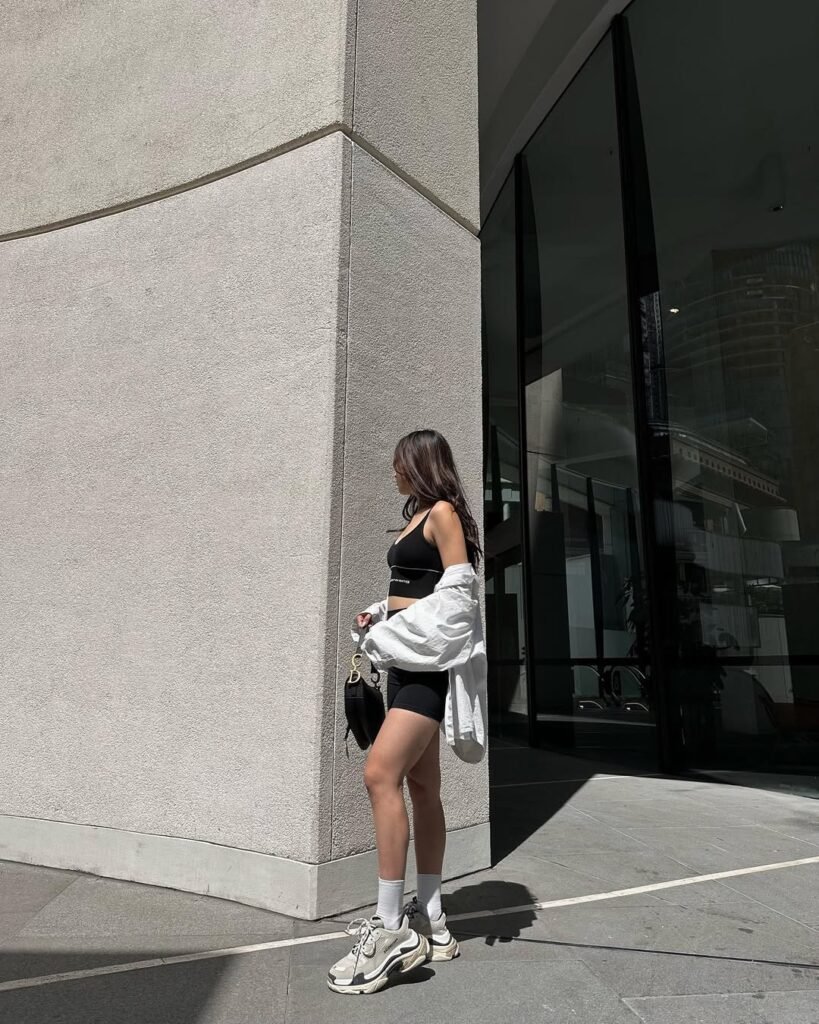 Woman wearing black biker shorts and crop top with a white shirt draped over shoulders, standing by a concrete wall in chunky white sneakers and carrying a black bag.