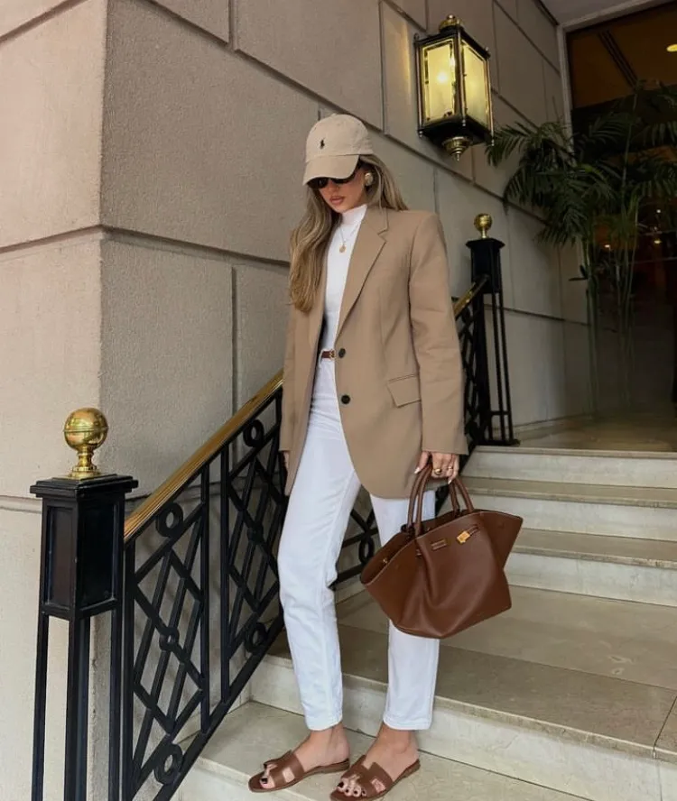 Woman wearing a camel blazer over a white top and white pants, accessorized with a tan handbag and sandals, standing on stairs. Blazer Outfits.