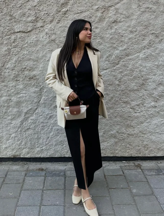 Woman posing in a cream blazer over a black dress with a front slit, holding a small handbag and wearing cream flats, against a textured wall. Blazer Outfits.

