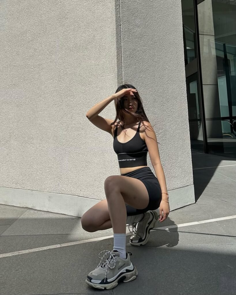A woman wearing a black crop top and black biker shorts is crouching outdoors. She has white socks and chunky white sneakers. She is shielding her eyes from the sun with one hand and has long dark hair flowing freely.