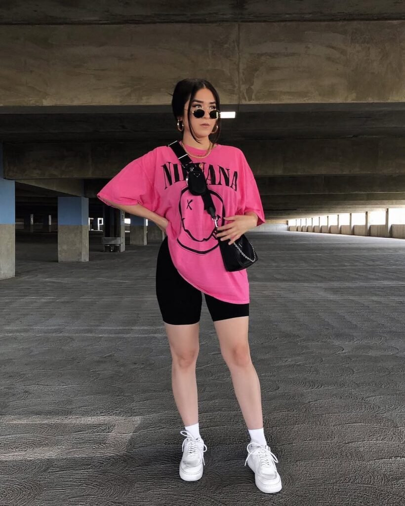 A woman is standing in a parking garage wearing a bright pink oversized Nirvana t-shirt and black biker shorts. She accessorizes with layered gold necklaces, hoop earrings, and a black crossbody bag. She completes the look with white socks and white sneakers.