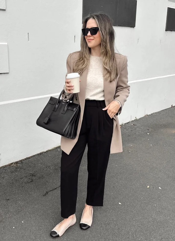 Woman in a beige blazer over a beige sweater and black pants, carrying a large black handbag and wearing black sunglasses, standing on a sidewalk. Blazer Outfits.