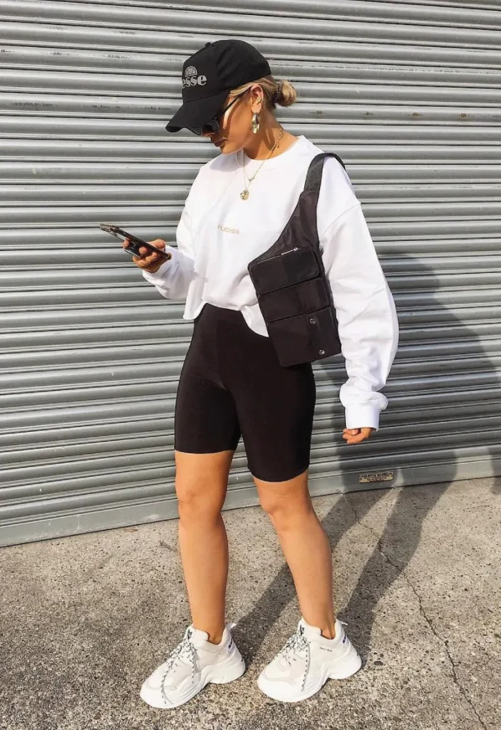 Casual biker shorts outfit featuring black biker shorts paired with a white oversized sweatshirt, black crossbody bag, and a black cap.