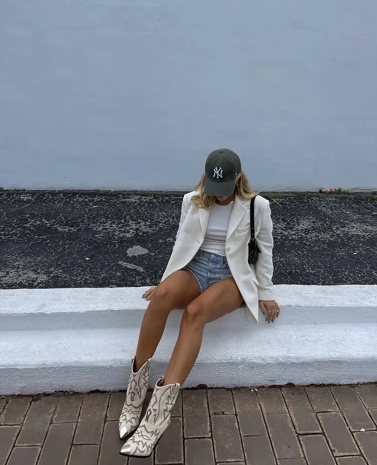 Woman sitting outdoors in a white blazer over a white tank top and denim shorts, paired with white cowboy boots and a green baseball cap. Blazer Outfits.