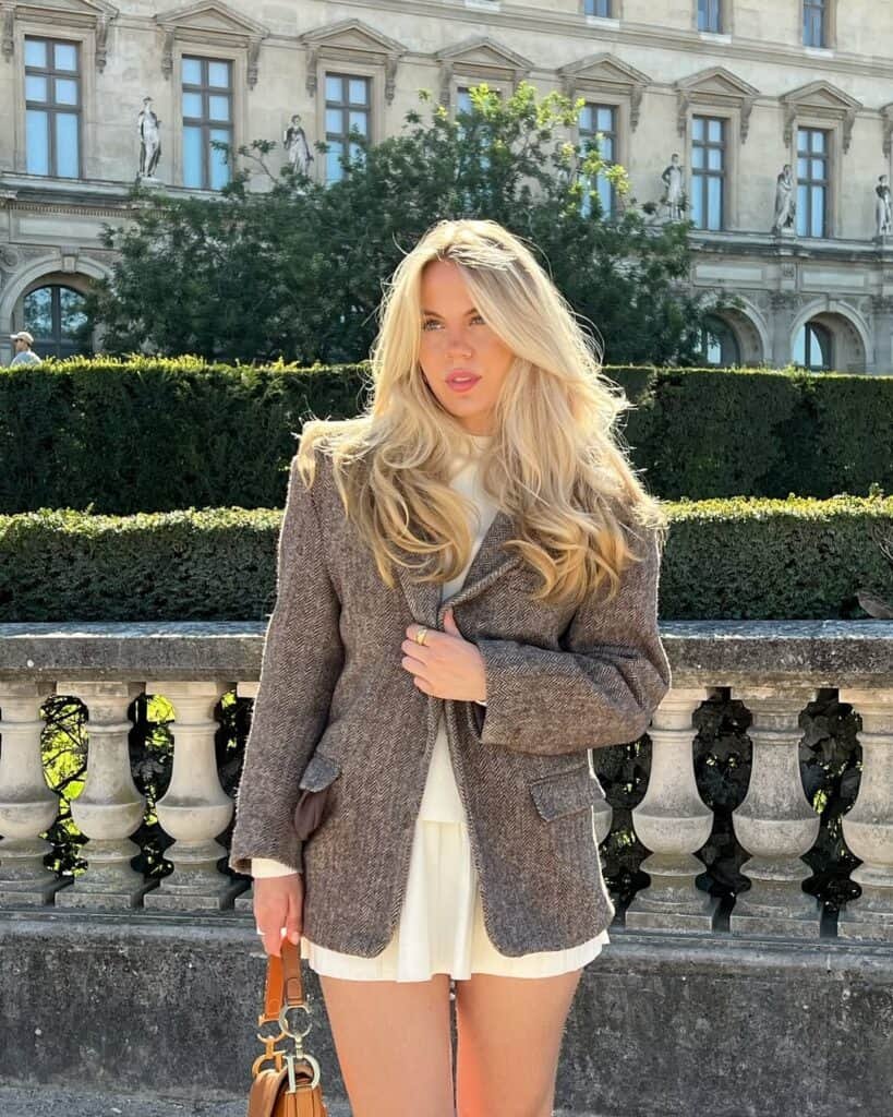 Woman wearing a brown textured blazer over a white dress, standing outdoors near a stone balustrade, showcasing elegant blazer outfits.
