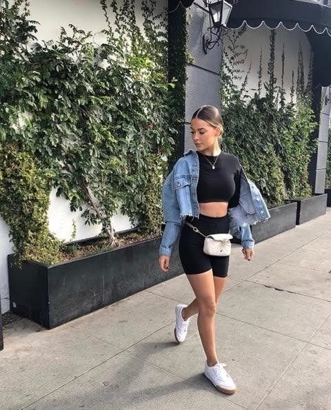 Woman walking on the street wearing black biker shorts outfit paired with a black crop top, light blue denim jacket, white sneakers, and a white waist bag. Stylish Biker Shorts outfits.