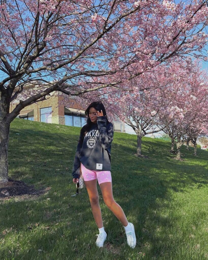 A woman posing outdoors under blooming trees in pink biker shorts, a dark oversized sweatshirt, and white sneakers – Biker Shorts outfits.