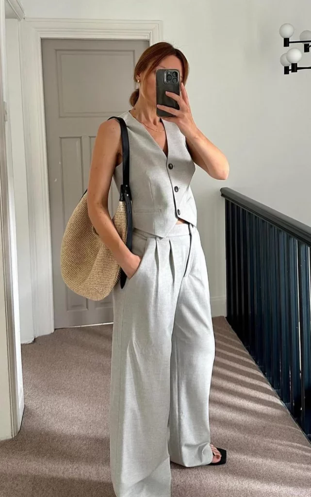 Woman wearing a sleeveless gray vest and wide-leg trousers with a straw tote bag, showcasing a refined and modern take on modest summer outfits.