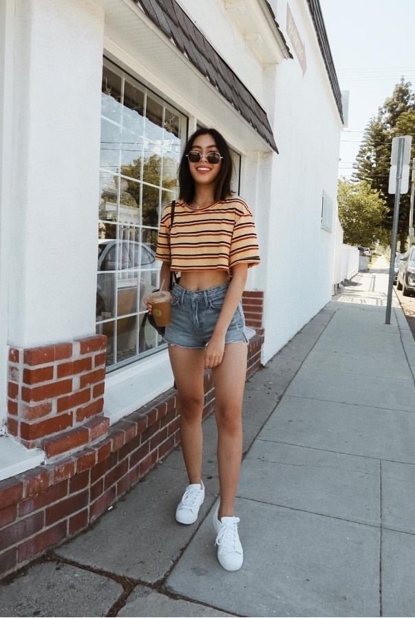 A young woman in a striped crop top and denim shorts paired with white sneakers, holding an iced drink—perfect example of casual Summer Outfits.