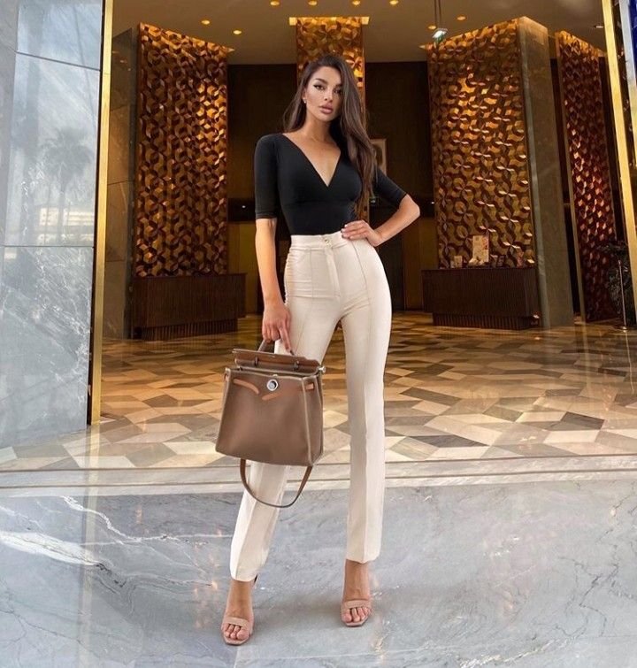 A woman in a chic black top and high-waisted cream trousers holding a tan structured handbag — look expensive outfits.