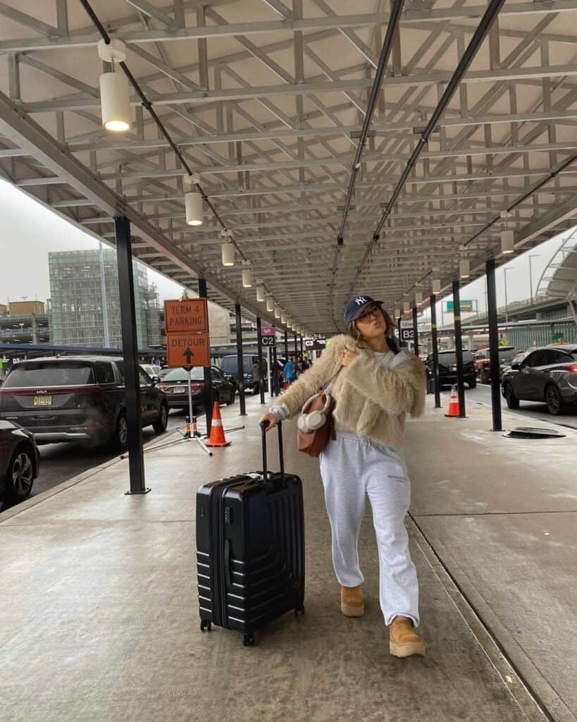 Cute Airport Outfits: Cozy traveler in grey sweatpants, fuzzy beige sweater, tan UGG boots, and a navy NY cap, walking outside an airport terminal with a black suitcase.