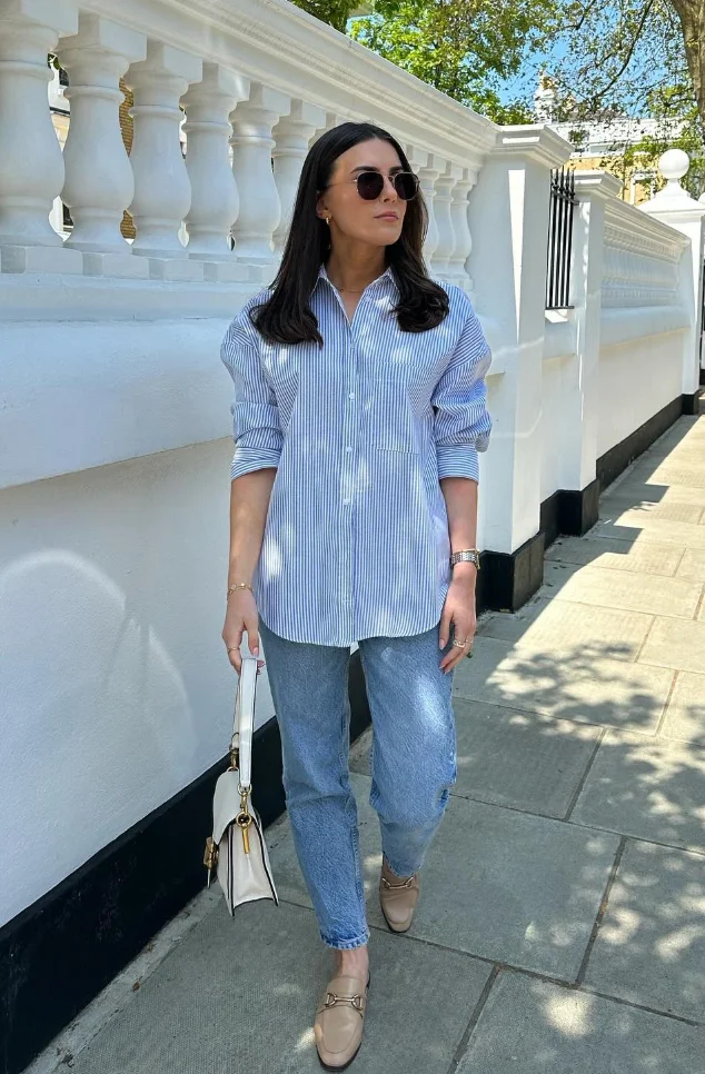  Woman in a loose-fitting blue striped shirt and straight-leg jeans with neutral loafers, showcasing a simple yet stylish modest summer outfit.