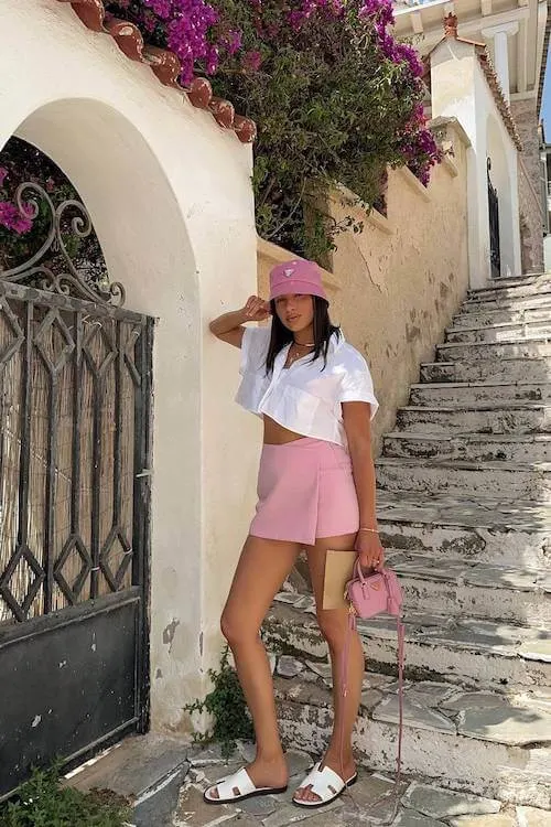 Playful Summer Vacation Outfit with a pink bucket hat, white cropped shirt, and pastel pink shorts, styled with white sandals and mini bag.