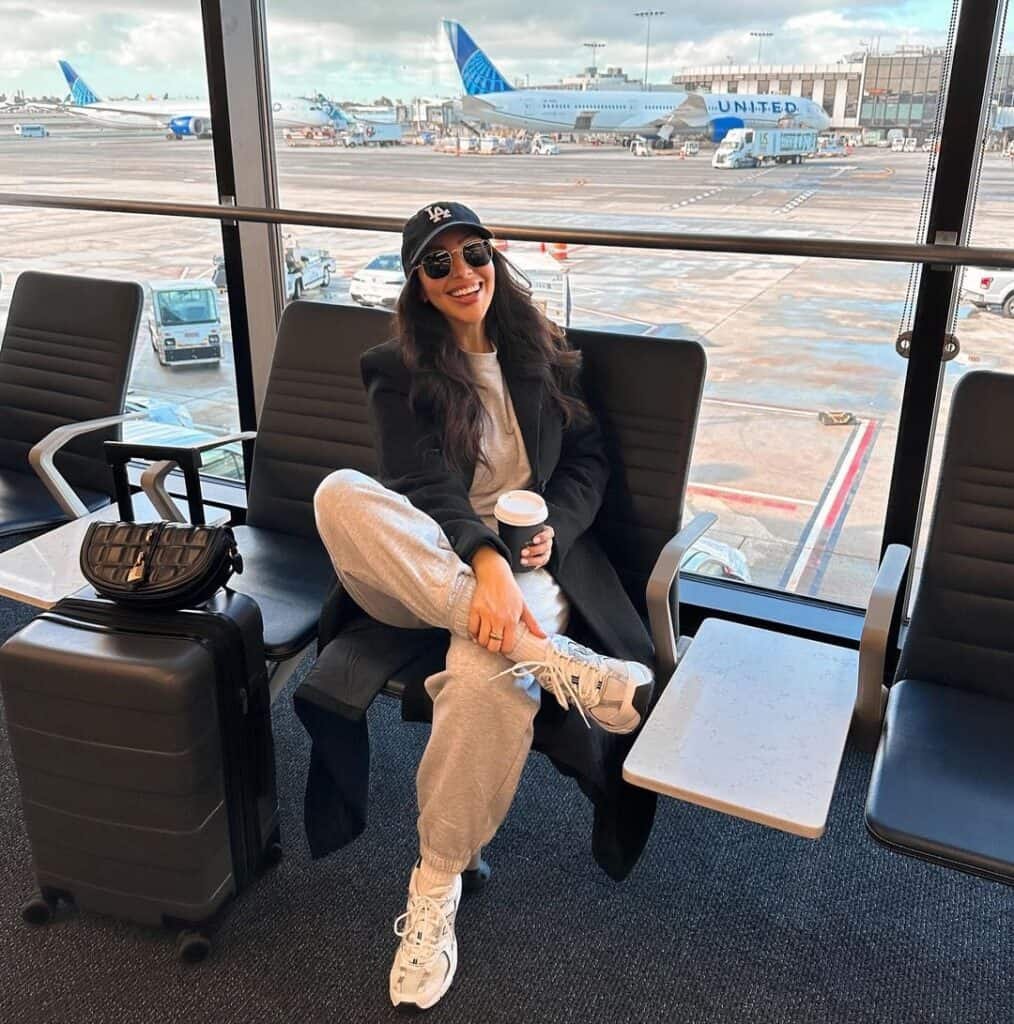 Cute Airport Outfits: Woman lounging in grey joggers, white sneakers, black coat, and a black cap, smiling with a coffee cup at the gate with planes visible through the window.