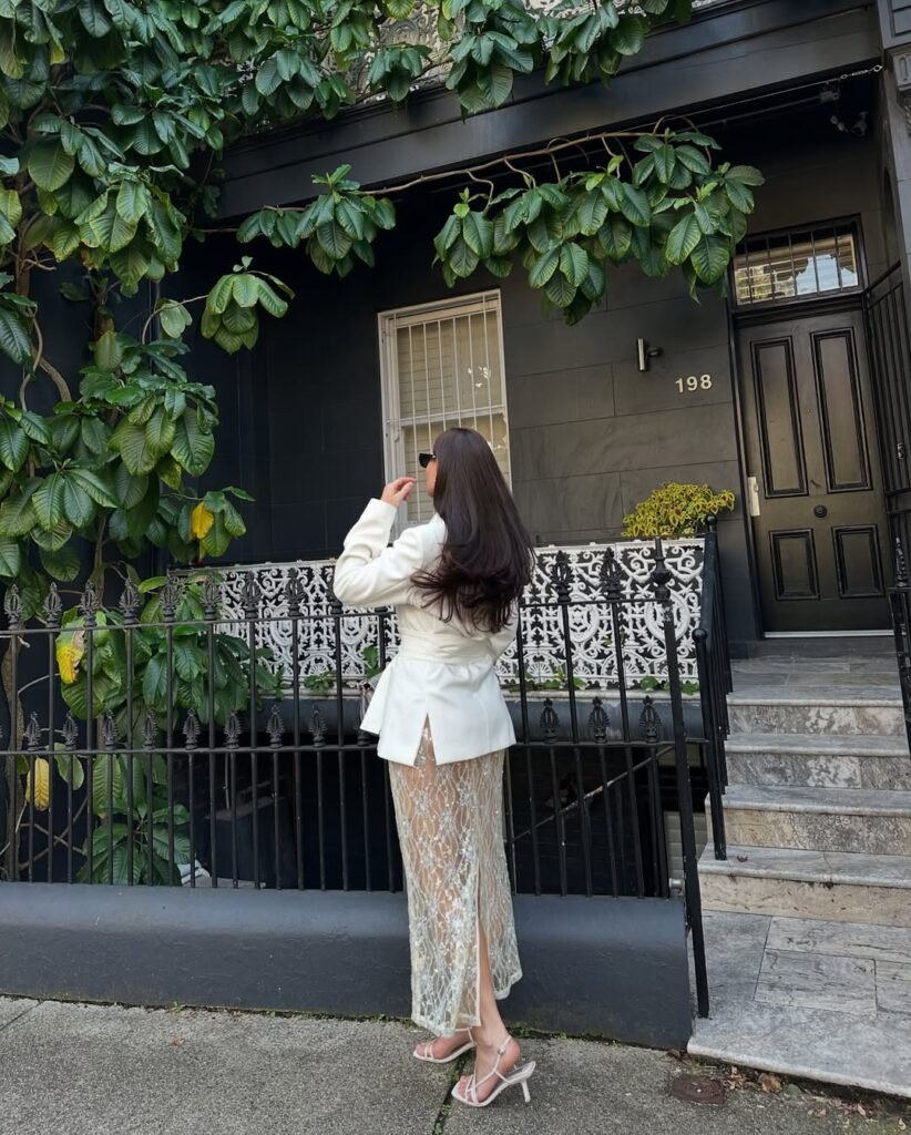 A woman in a cream structured blazer and an intricate lace midi skirt with high heels, standing in front of a black townhome – sophisticated and refined old money spring look.