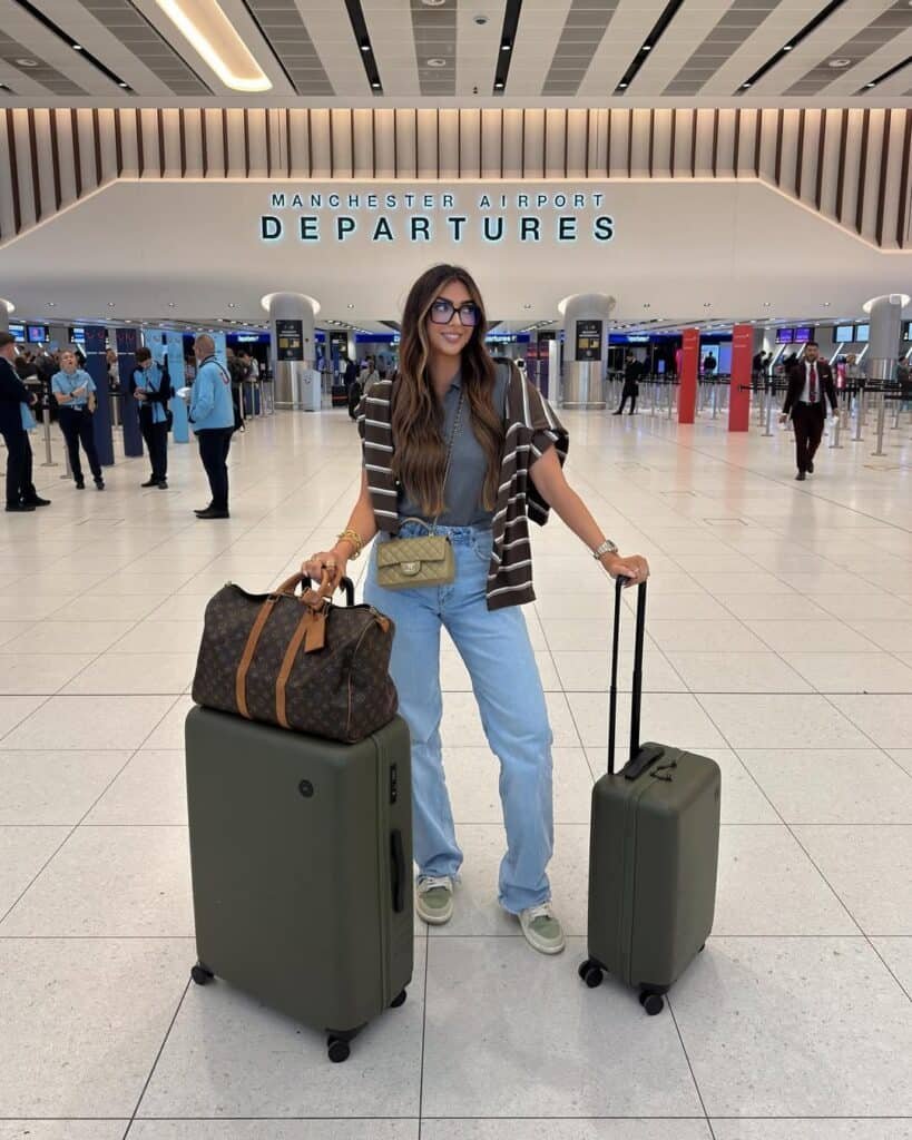 Cute Airport Outfits: Trendy traveler in blue jeans, a grey tee, striped cardigan, and designer crossbody bag at Manchester Airport, with olive green suitcases and a Louis Vuitton duffel.