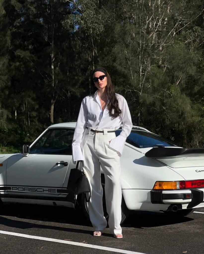 A woman wearing a crisp white shirt tucked into high-waisted white trousers, accessorized with black sunglasses and a black handbag, standing by a vintage Porsche – elevated old money spring chic.