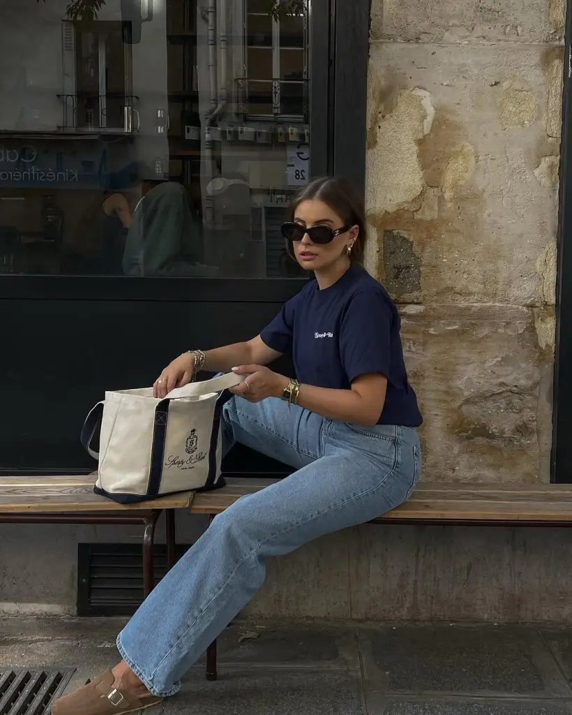 Relaxed Casual Summer Outfits with a navy tee and classic light blue jeans, paired with beige Birkenstock-style sandals and black square sunglasses while sitting on a bench with a large tote.