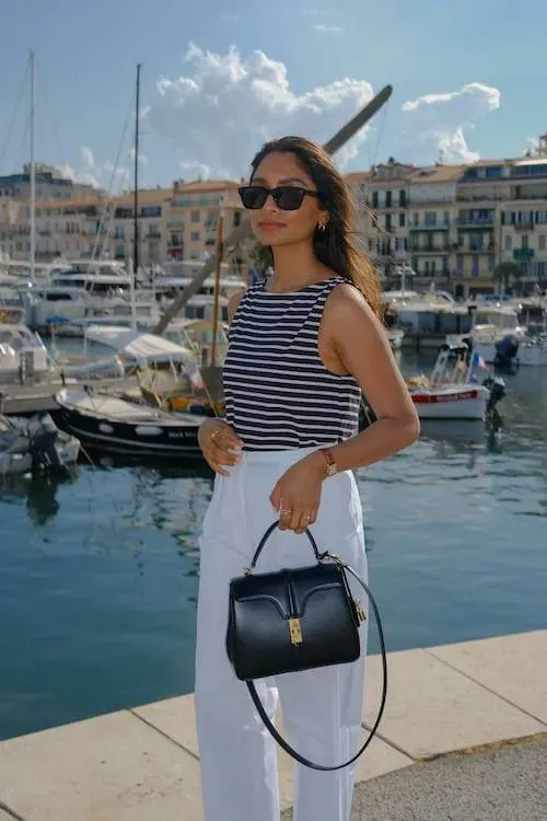 Classic Summer Vacation Outfit with a navy and white striped sleeveless top, white high-waisted pants, and a structured black handbag by the marina.