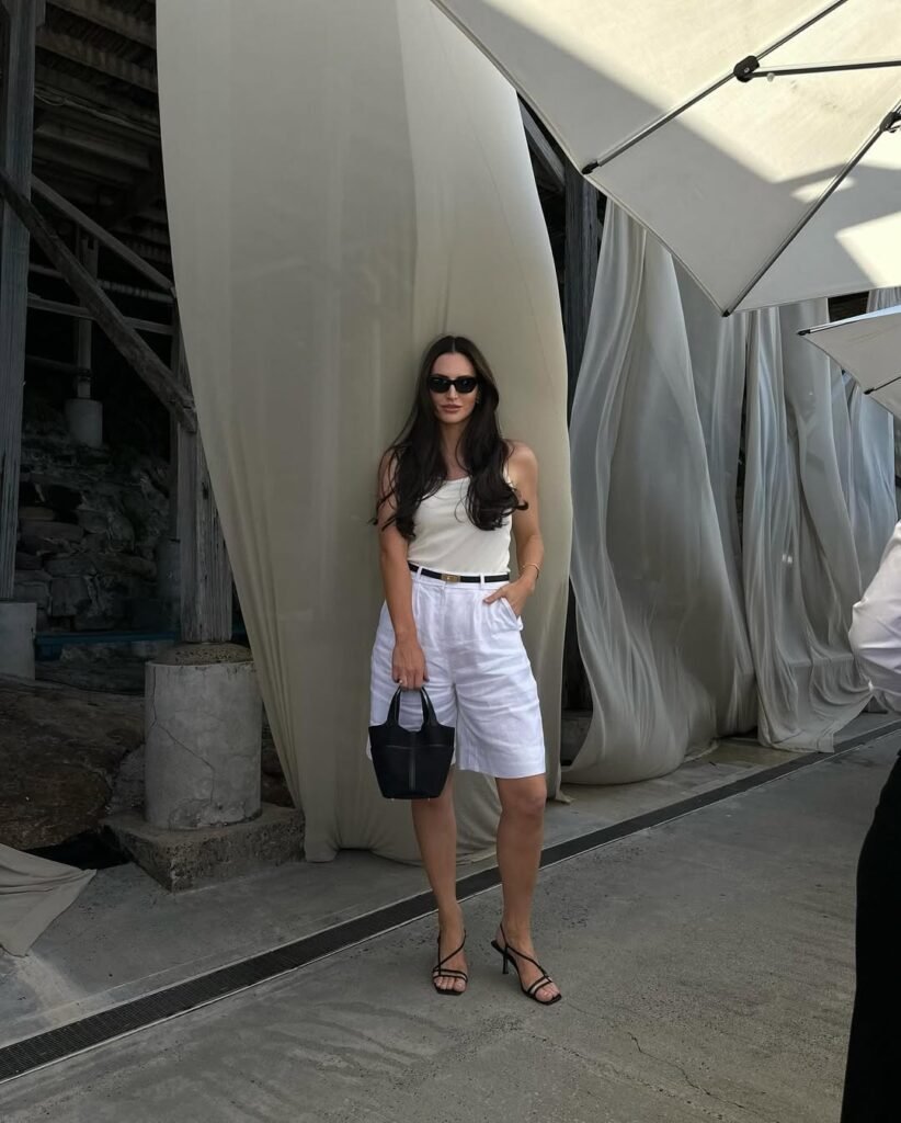 A woman dressed in a sleeveless white top and loose white Bermuda shorts, styled with black strappy heels and a minimalist bag – understated and breezy old money spring outfit.