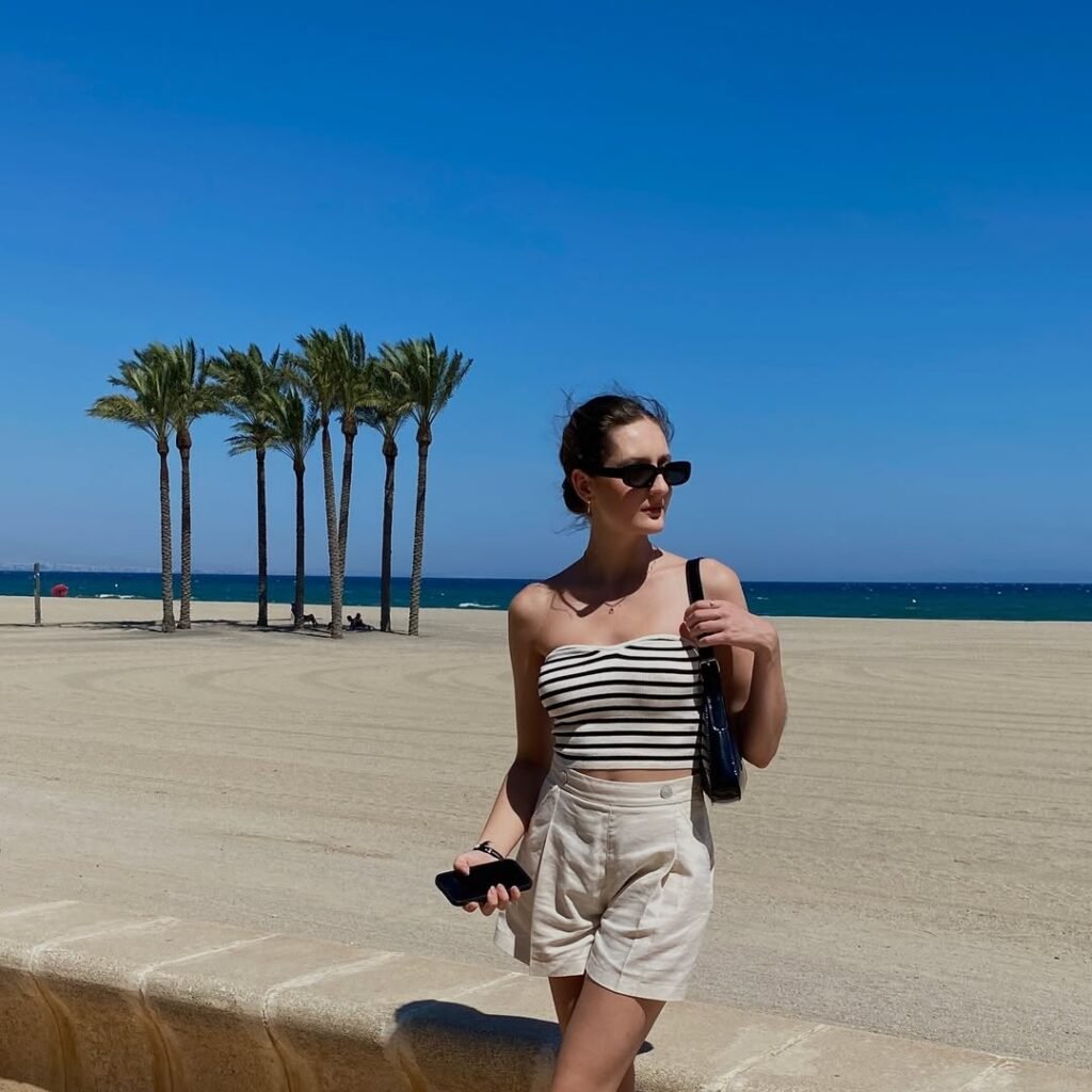 A woman at the beach in a striped bandeau top and high-waisted linen shorts with cat-eye sunglasses – a coastal-inspired old money spring outfit.